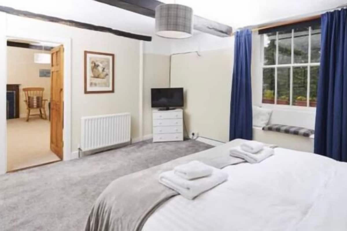 Une chambre à coucher comprenant un lit, un téléviseur, un radiateur, une fenêtre avec des rideaux et une porte donnant sur une autre pièce.