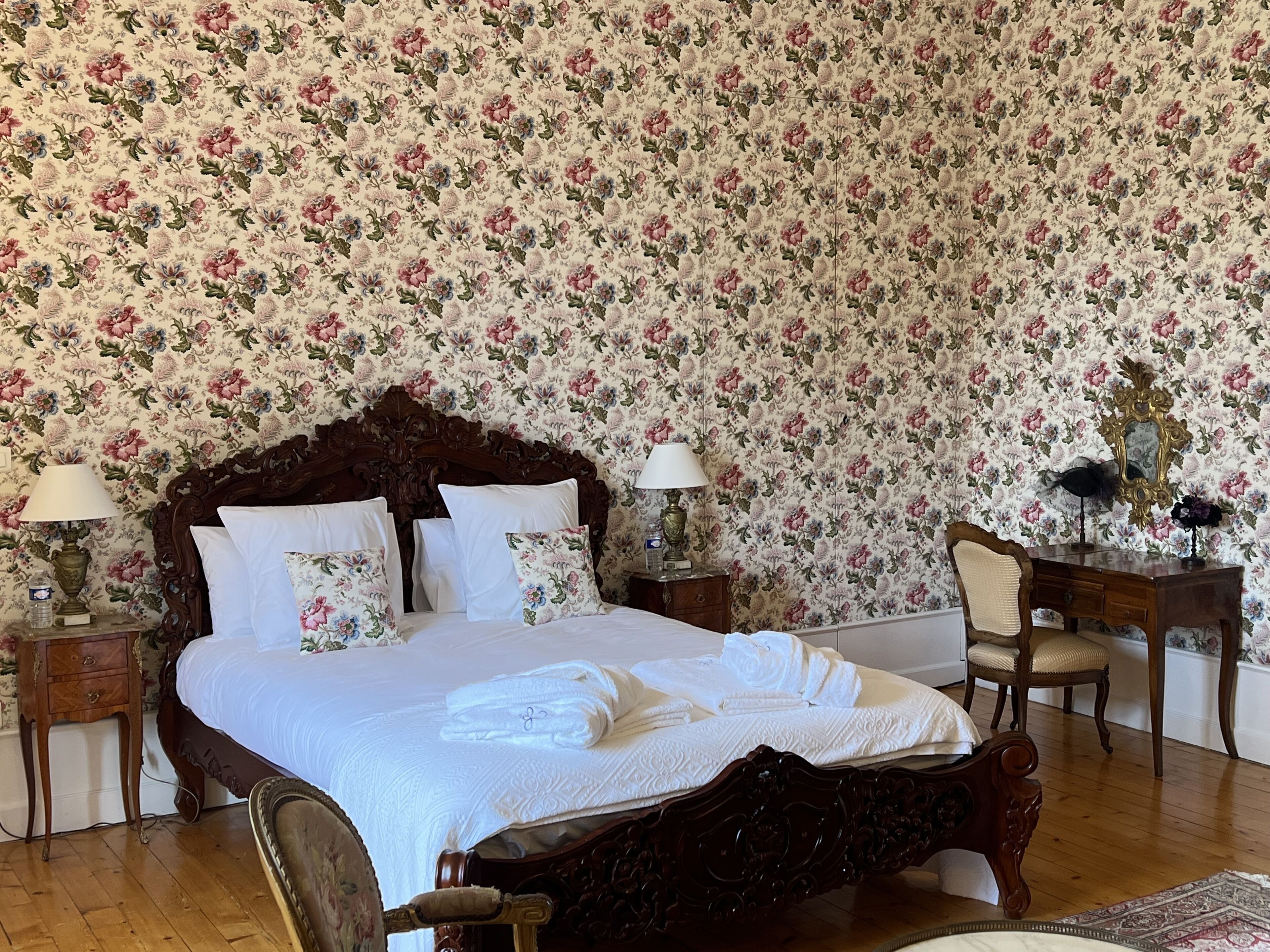A bedroom with a floral wallpaper, a wooden bed with white linens, two bedside tables with lamps, a wooden chair, and a dresser.