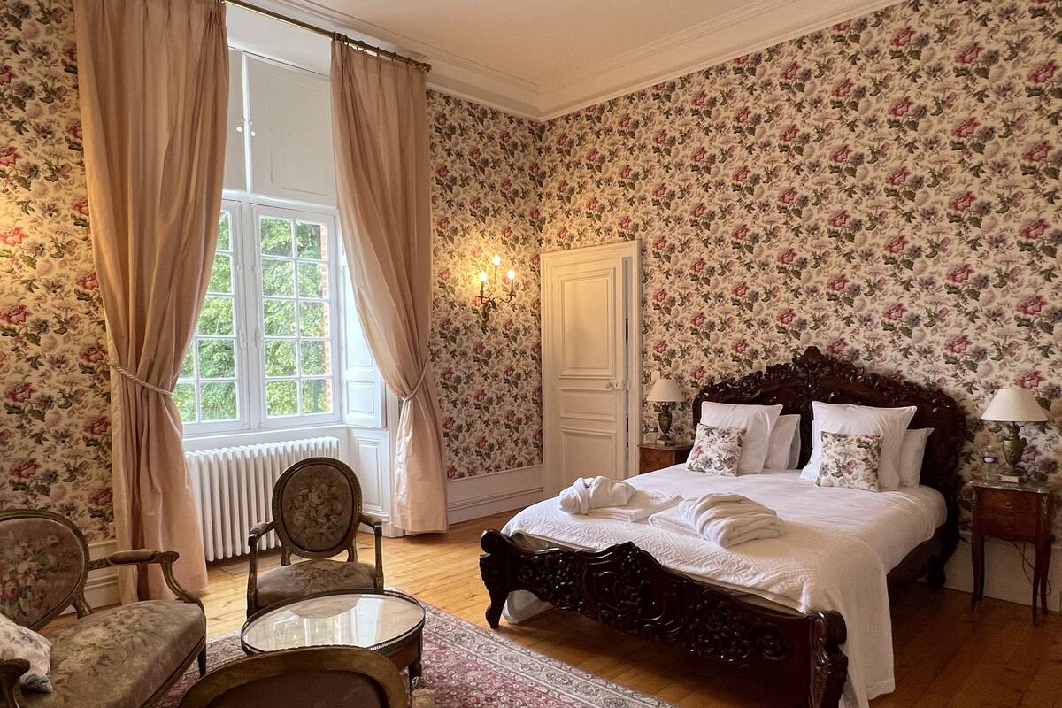 A bedroom with a large bed, floral wallpaper, a window with curtains, a radiator, and a wooden chair.