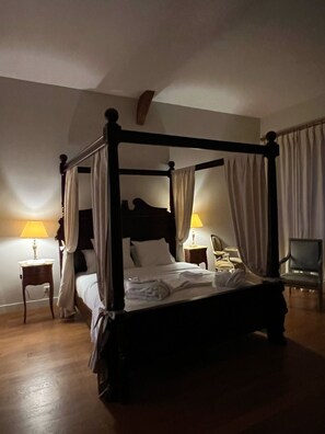 A four-poster bed with white linens, two bedside tables with lamps, and a chair in a dimly lit room with wooden flooring. - Château de Lauresse (Lombron)