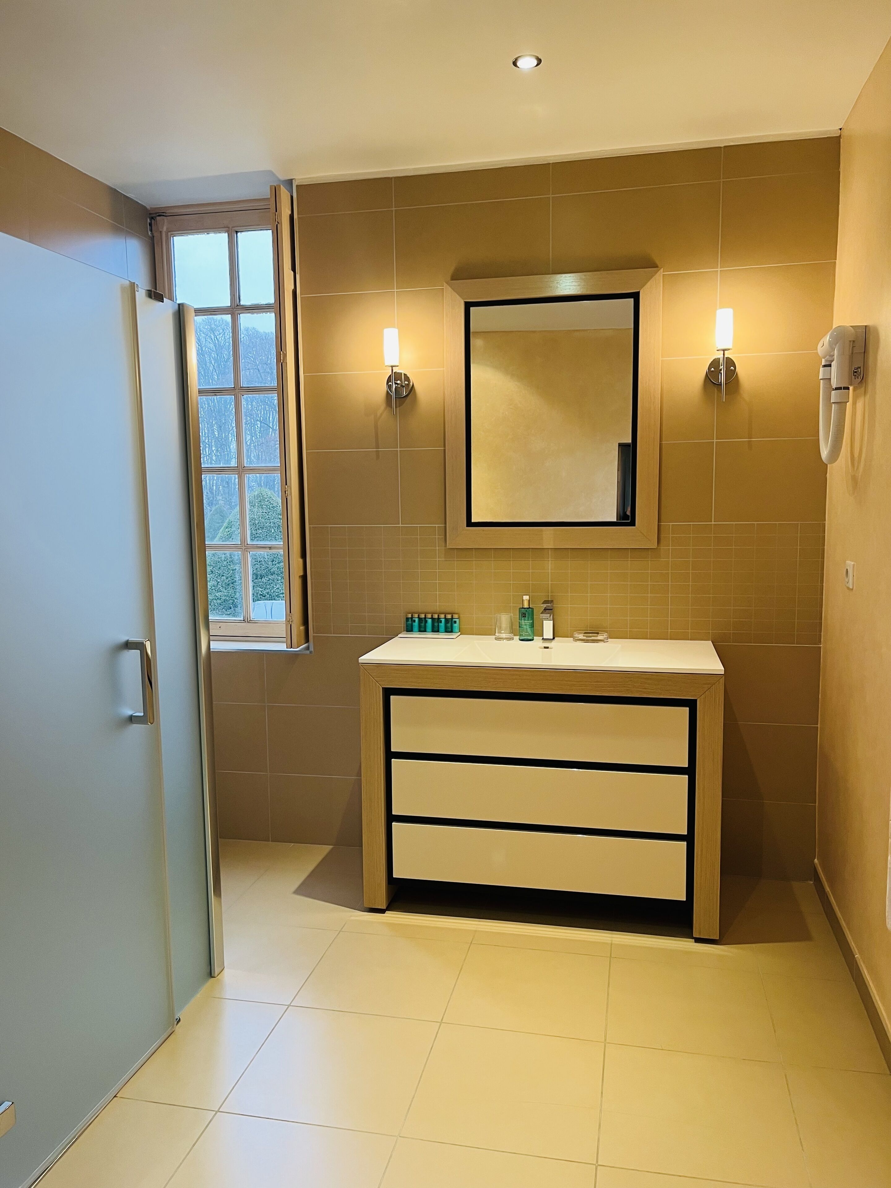 A bathroom with a beige tiled wall, a wooden vanity with a mirror, two wall-mounted lights, and a window with a view of trees.