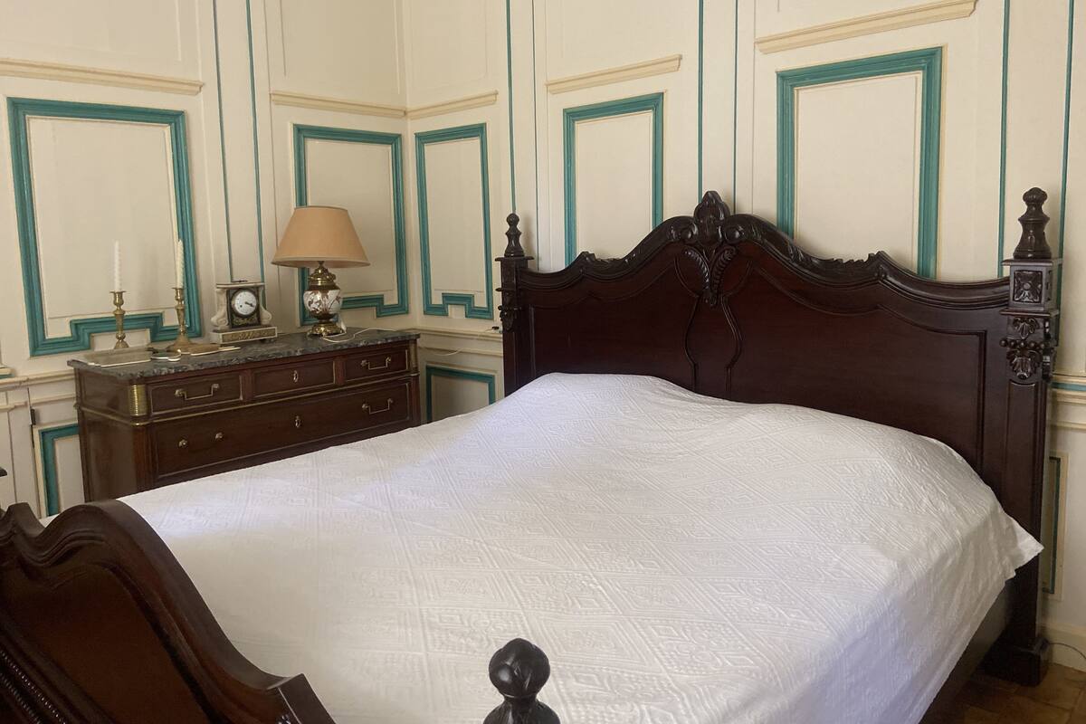 A neatly made bed with a white bedspread in a room with teal and cream wall paneling, a wooden dresser with drawers, and a small table with a lamp and a clock.