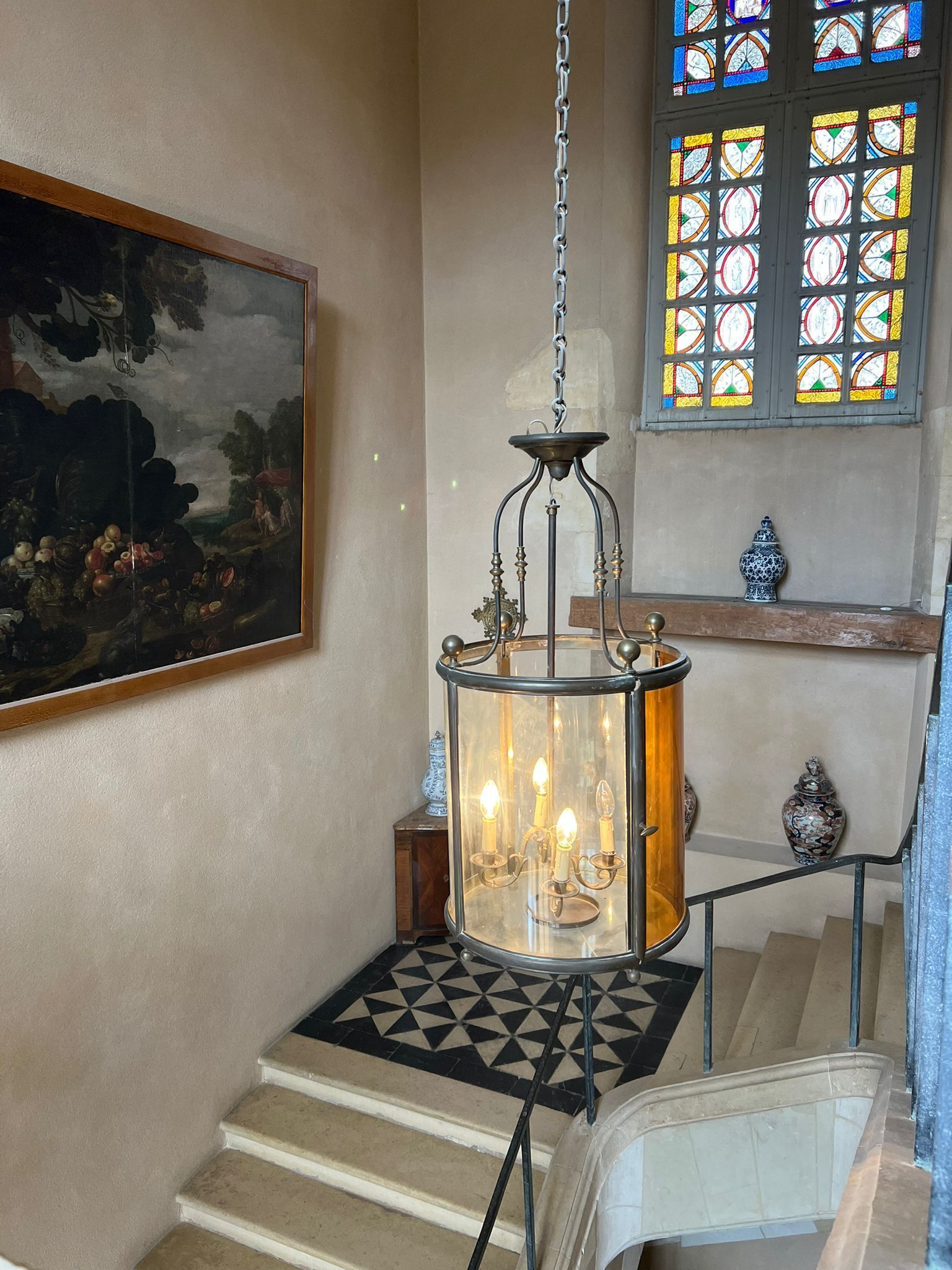 A staircase with a patterned carpet, a hanging lantern with candles, a painting on the wall, and a stained glass window.