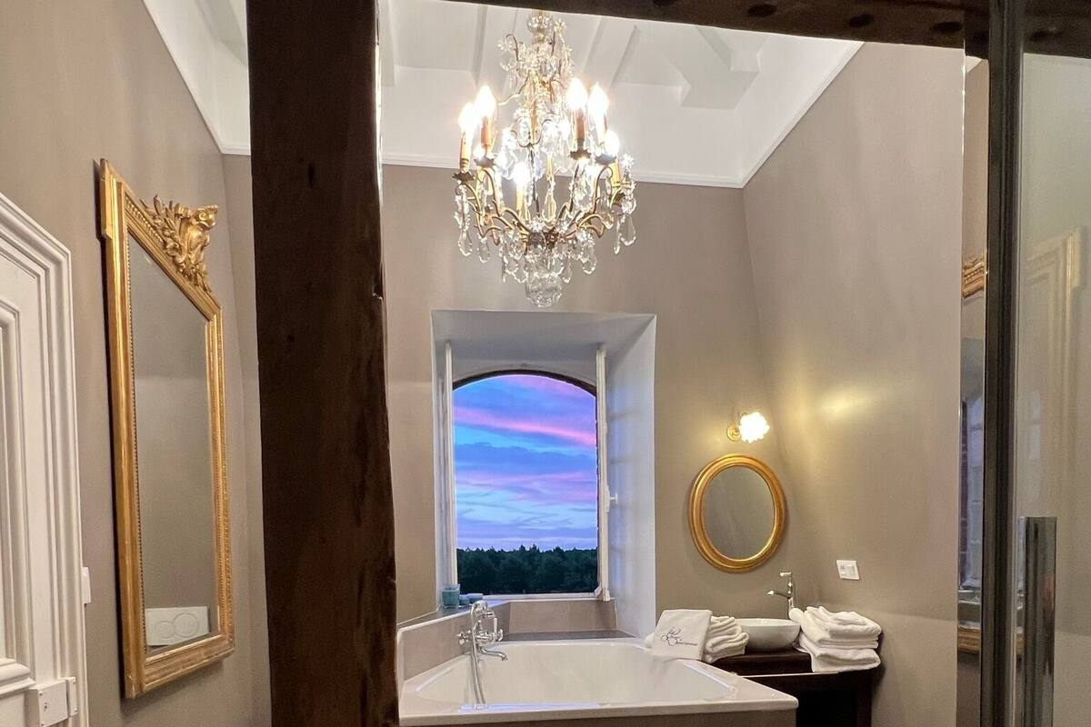 A modern bathroom with a freestanding bathtub, a chandelier, and a view of the sky through a window.