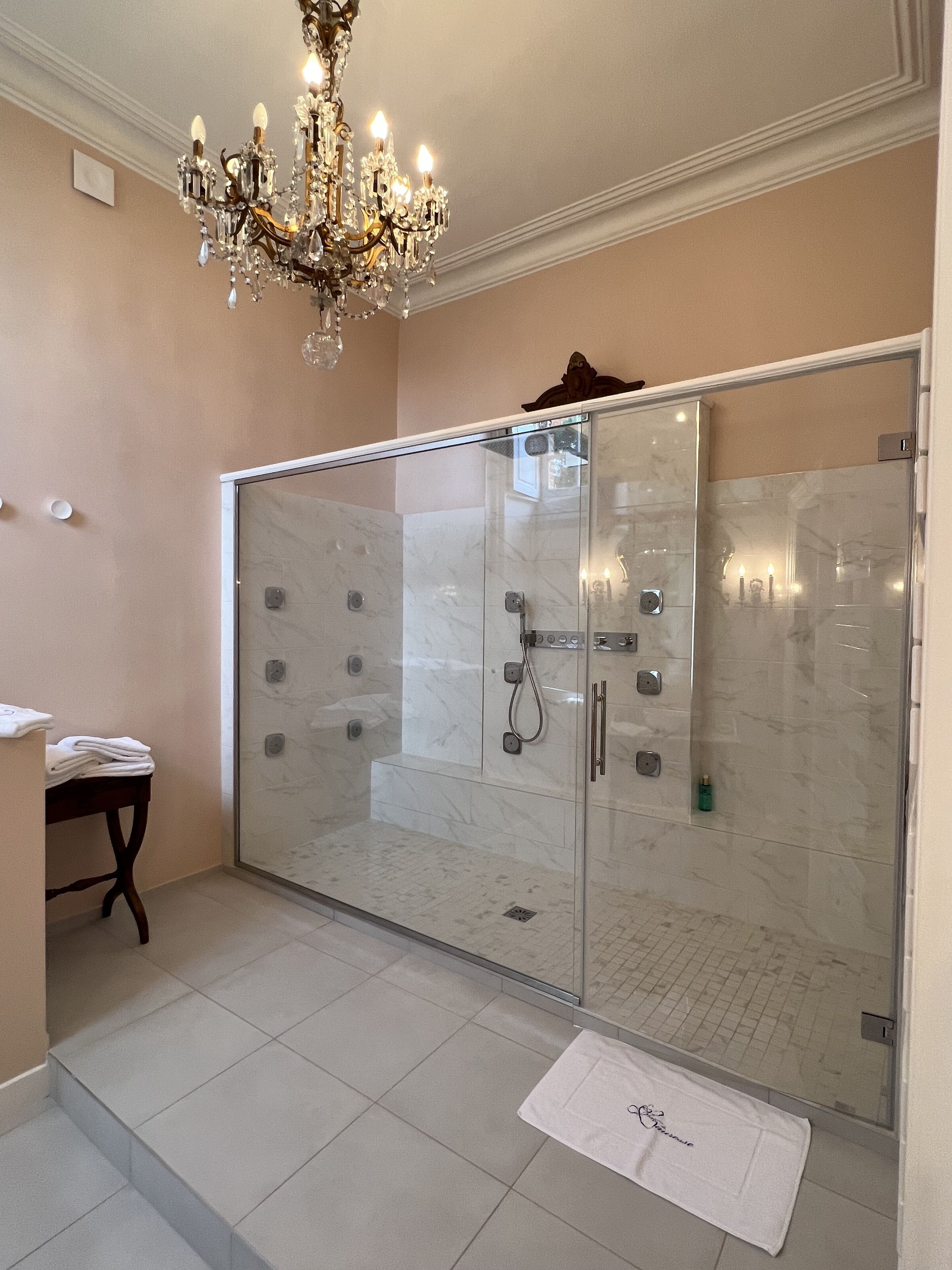 A bathroom with a glass shower enclosure, a marble-tiled floor, a white towel on the floor, and a chandelier.