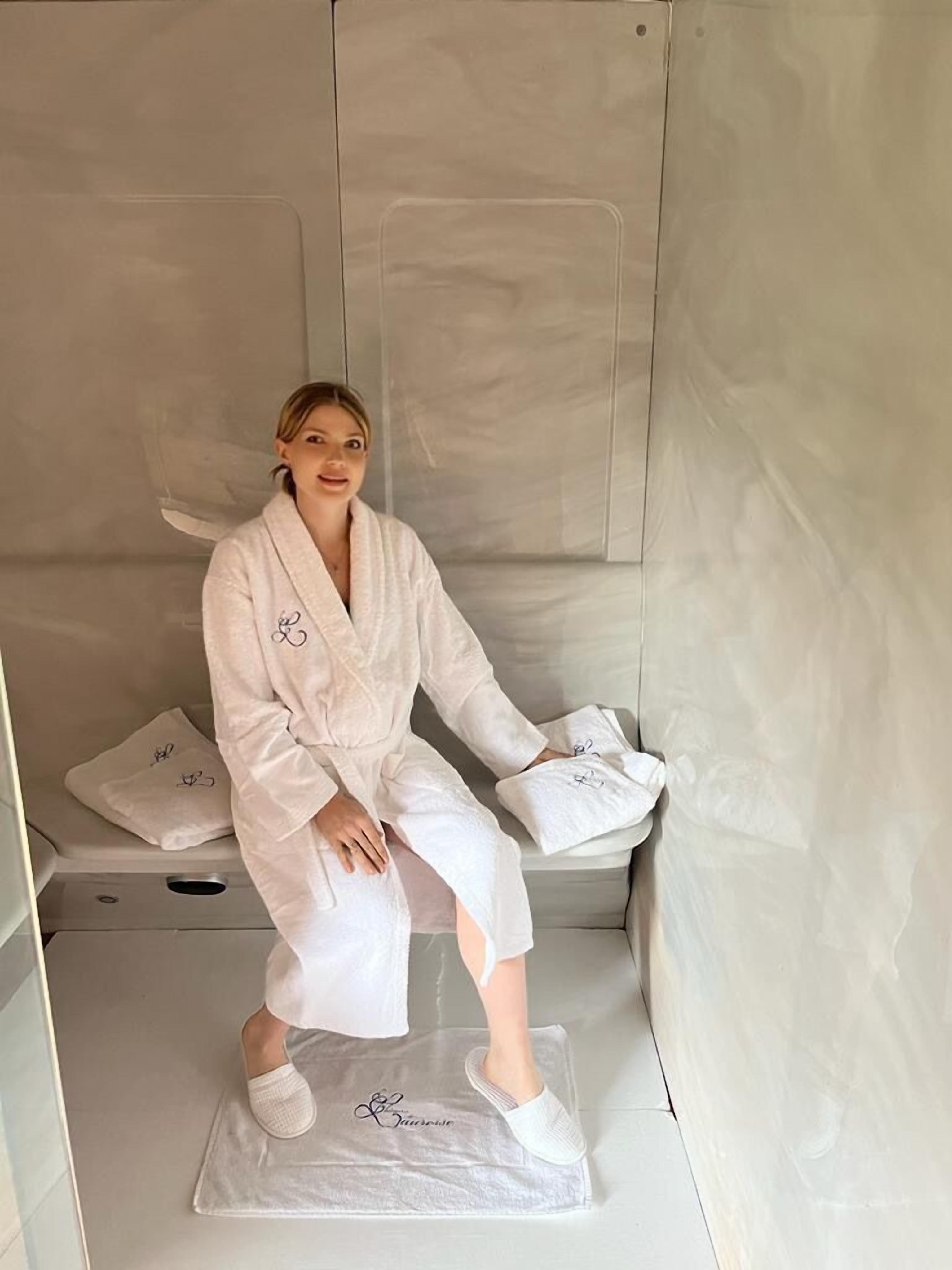 Une personne assise sur un banc, dans un cadre évoquant un spa, avec une serviette et des chaussons.