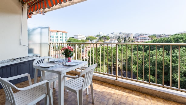 Un balcón con mesa y sillas, una parrilla y vistas a edificios y árboles.