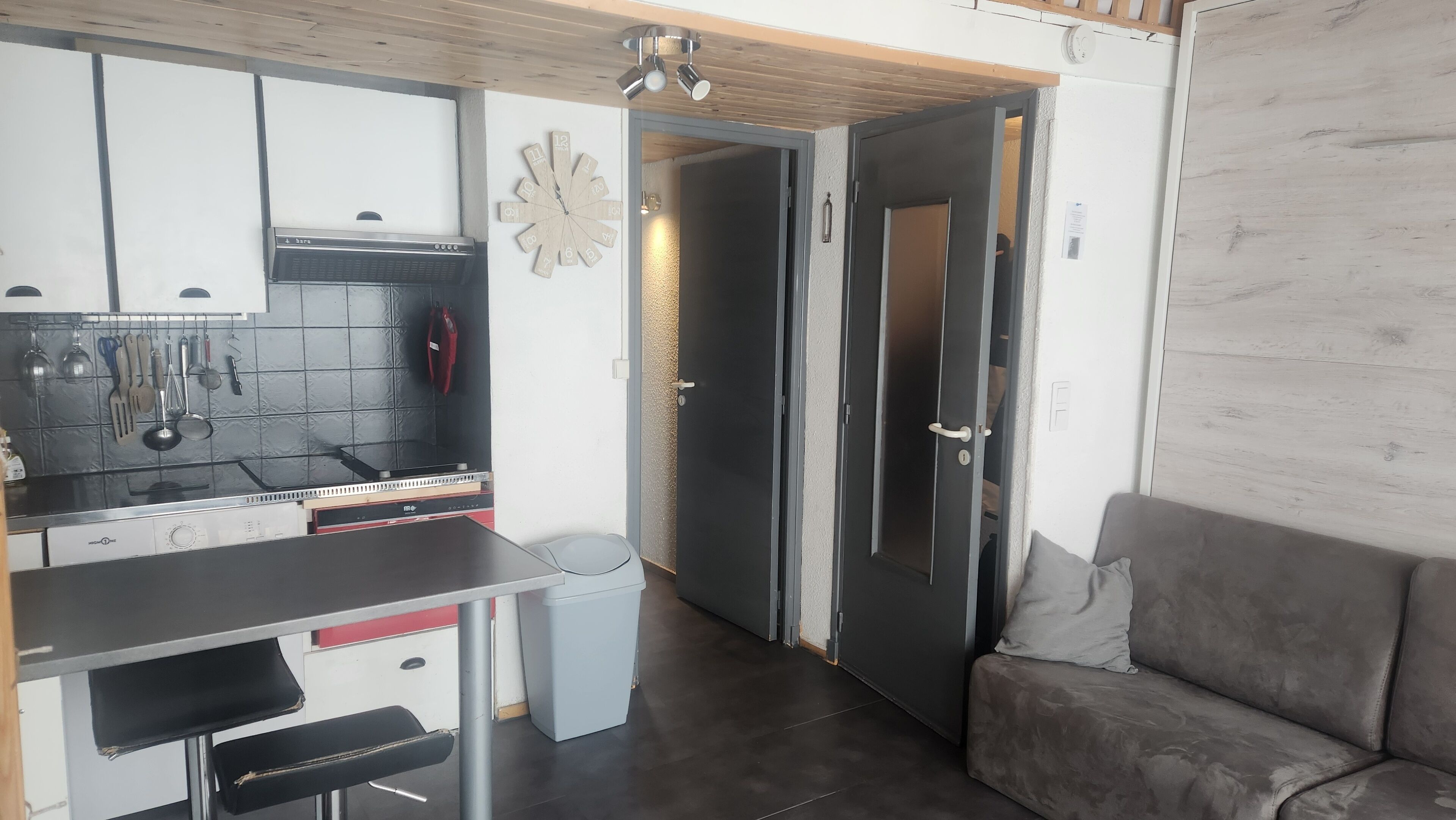 A compact kitchen with a stove, sink, and hanging utensils. A door leads to another room, and a couch is visible in the corner.