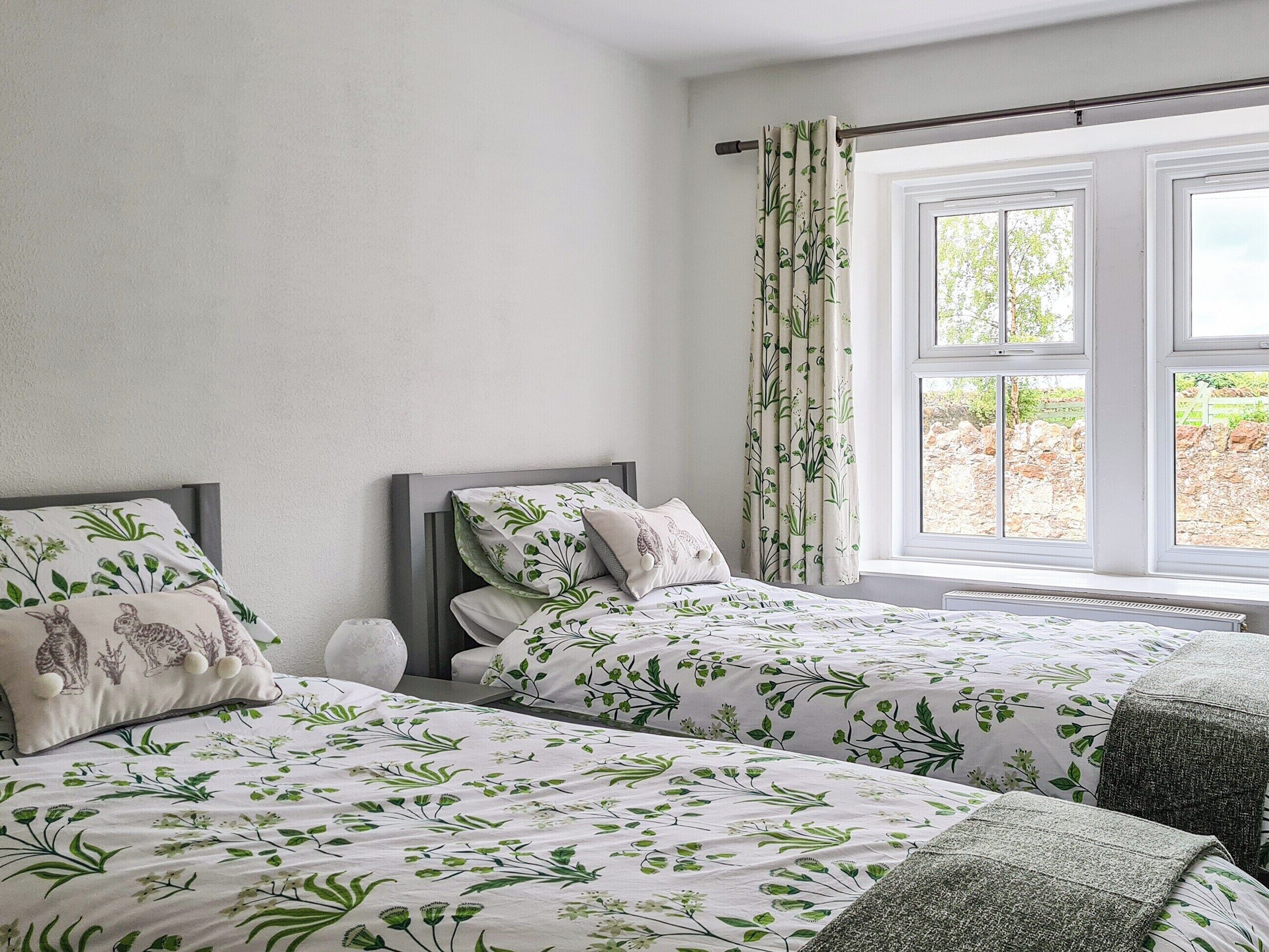 Une chambre avec deux lits, chacun agrémenté de draps à motifs floraux, une fenêtre avec des rideaux et une vue sur la verdure environnante.
