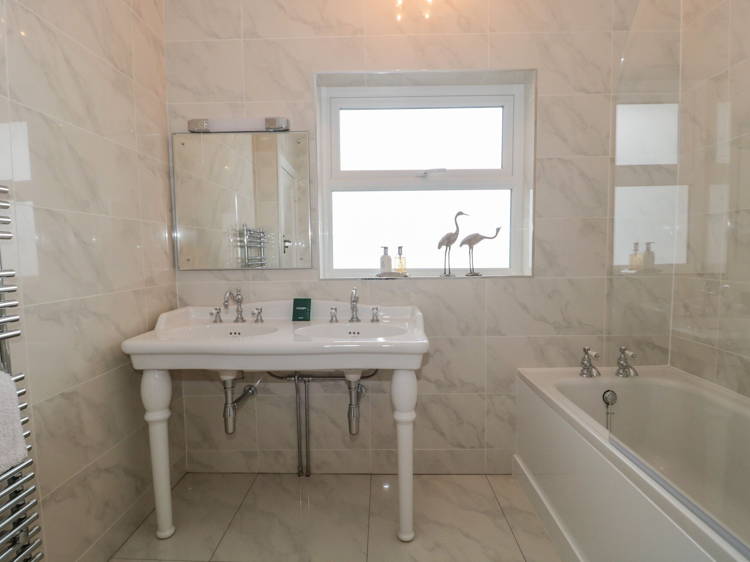 Une salle de bain avec un lavabo double, une baignoire, un miroir et une fenêtre ornée de deux figurines d’oiseaux décoratives.
