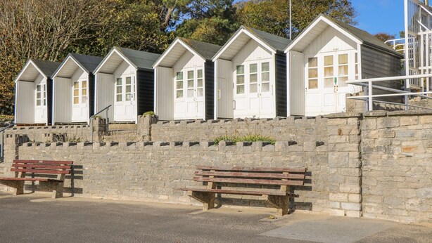 Uma fileira de cabanas brancas à beira-mar, com janelas, um muro de pedra e dois bancos de madeira.