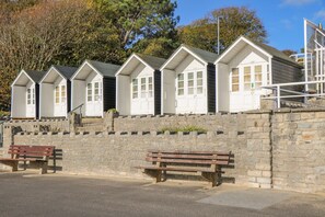 Uma fileira de cabanas brancas à beira-mar, com janelas, um muro de pedra e dois bancos de madeira.