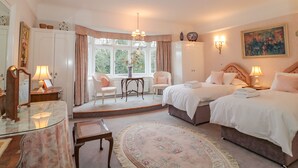 Quarto com duas camas, área de estar com cadeiras, mesa de vidro e uma janela grande com cortinas.