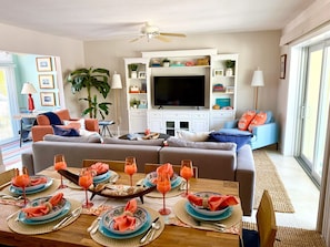 A modern living room with a sofa, a dining table set for a meal, and a television in the background. - Rum Point Island House #14, Waterfront, Beach, Pool, Bikes, Kayaks, Paddleboards (Rum Point, North Side)