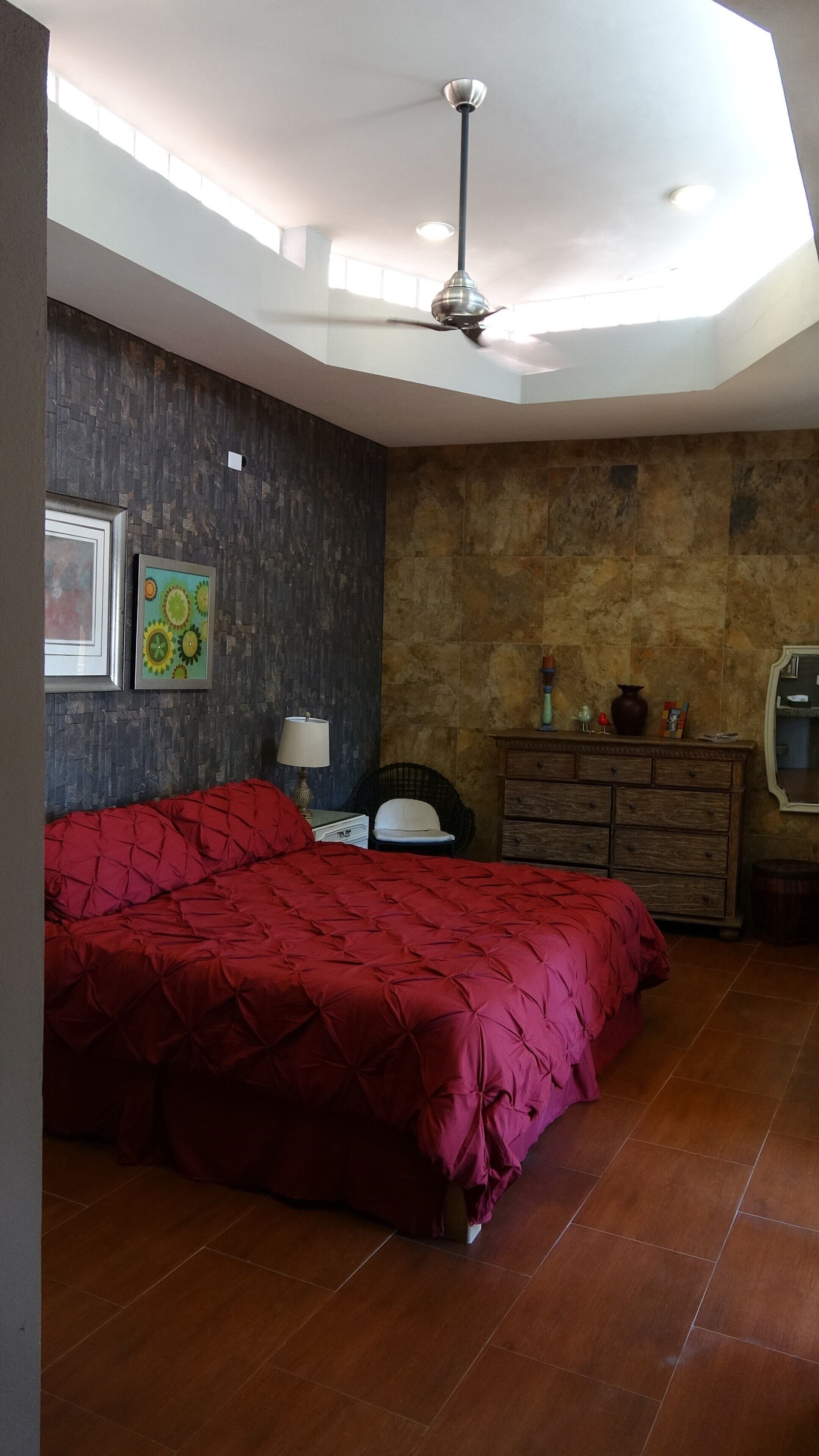 A bedroom with a large bed, a ceiling fan, a dresser, and a wall with a clock and artwork.