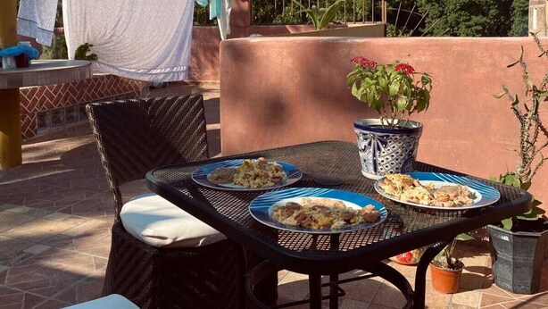 An outdoor patio with a table set for two, a wicker chair, and a view of a rocky mountain. - Casona 13 Flechas (Mazatlán)