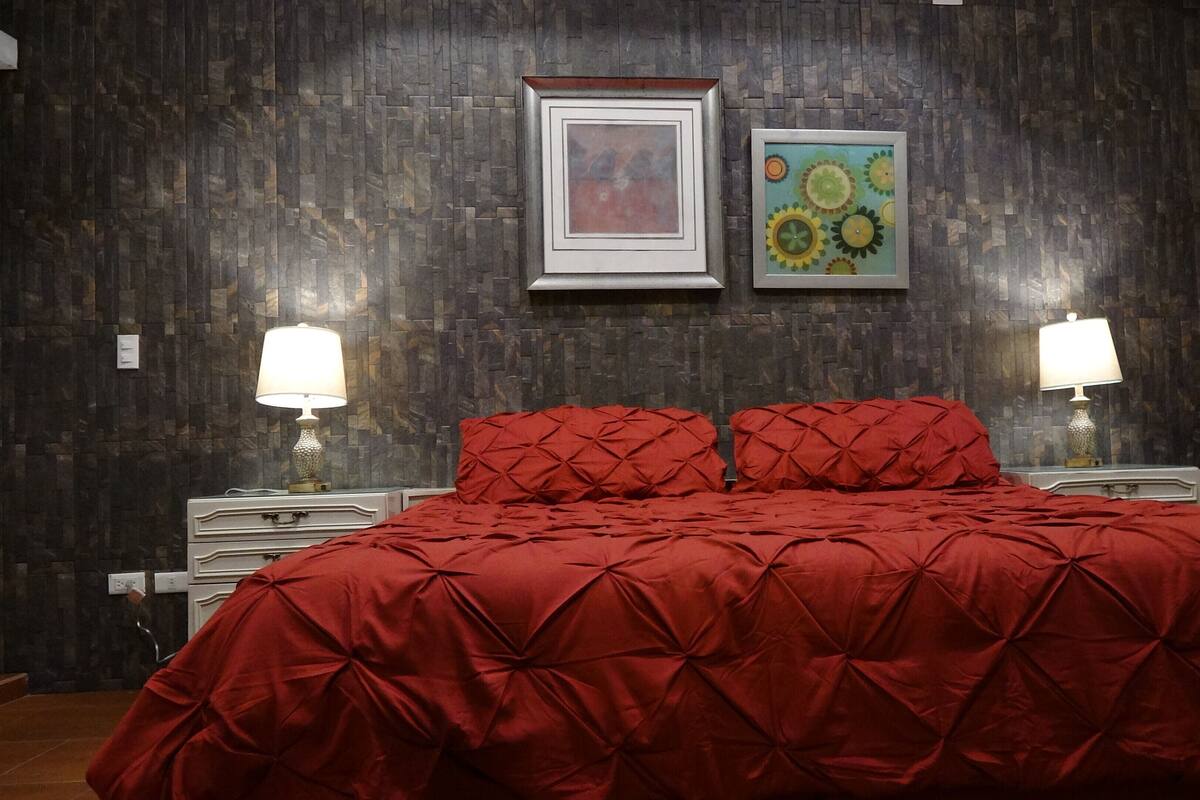 Quarto com parede de pedra escura, colcha vermelha, duas luminárias de cabeceira e duas obras de arte emolduradas.