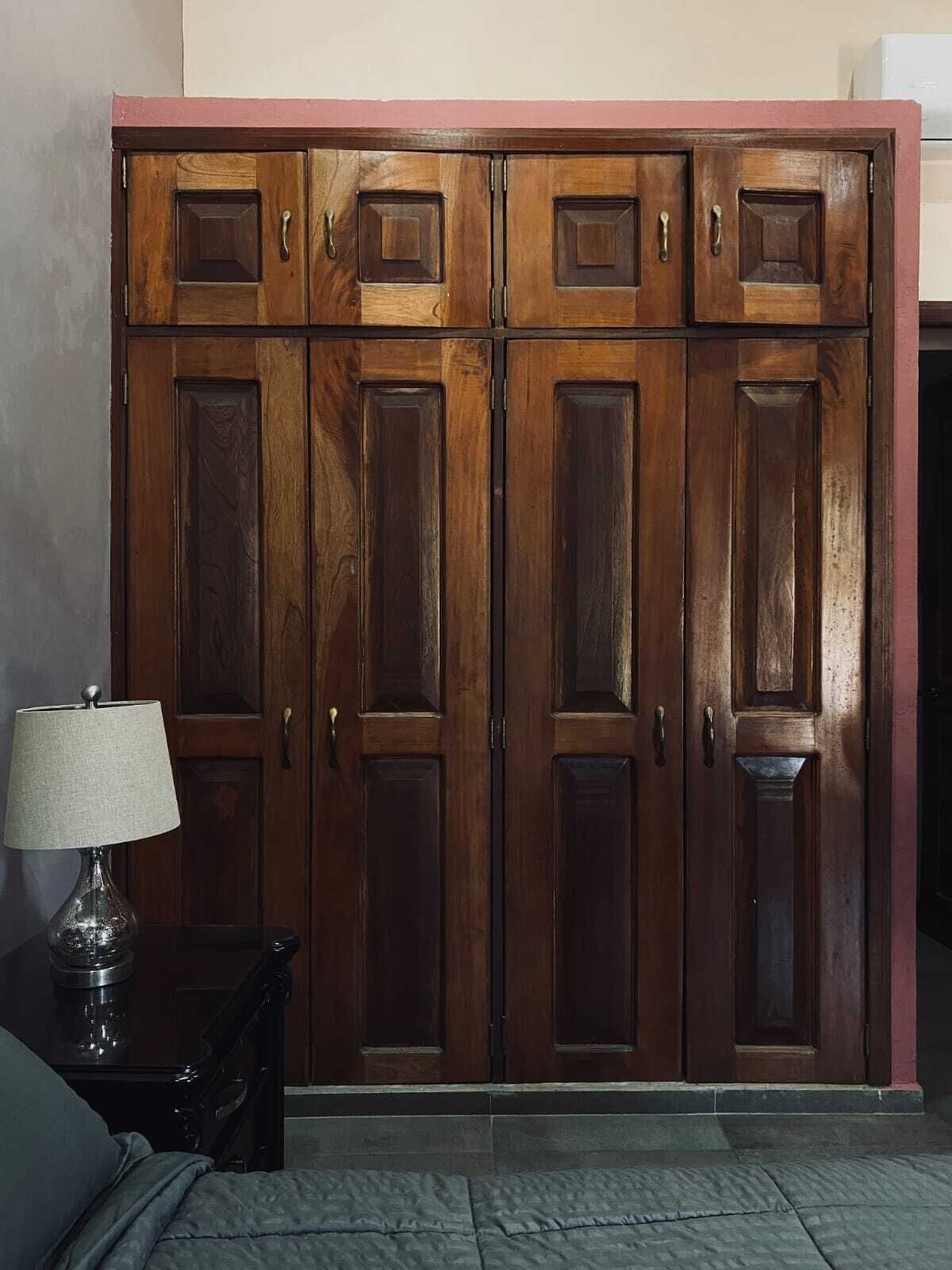 Een houten garderobe met meerdere deuren en handgrepen, een lamp op een bijzettafeltje, en een grijs vloerkleed.
