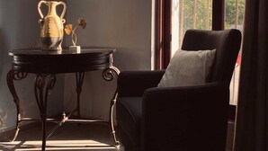 A room with a dark armchair, a small table with a vase, and a window with curtains. - Casona 13 Flechas (Mazatlán)