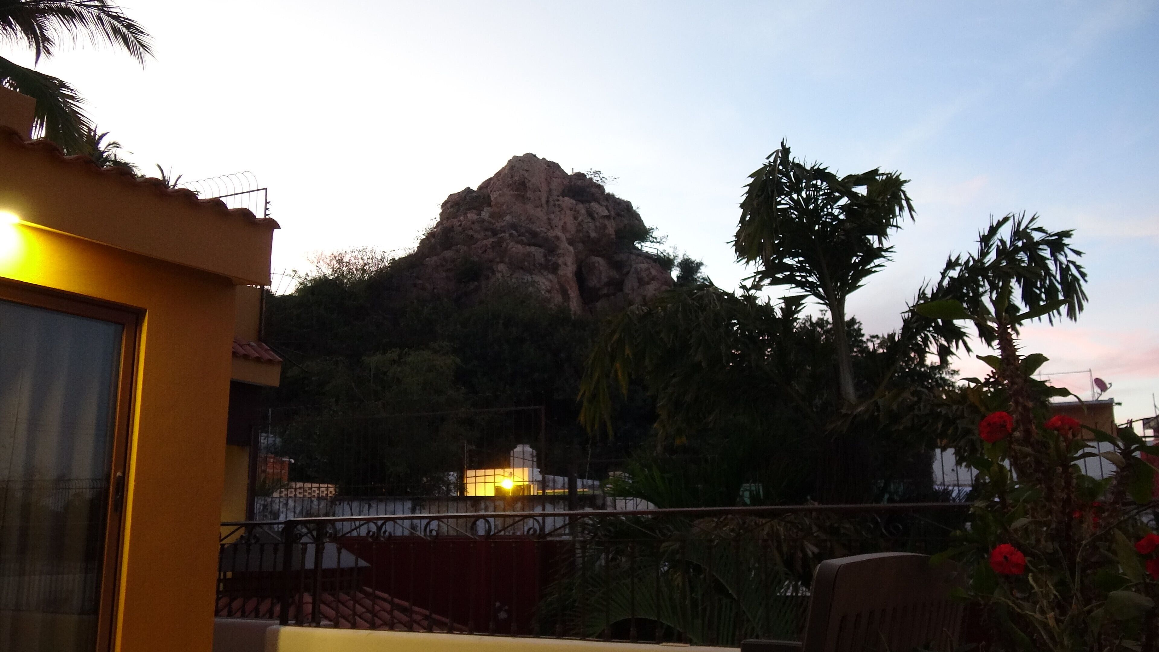 A balcony with a view of a rocky mountain, surrounded by greenery and a building with a yellow facade.