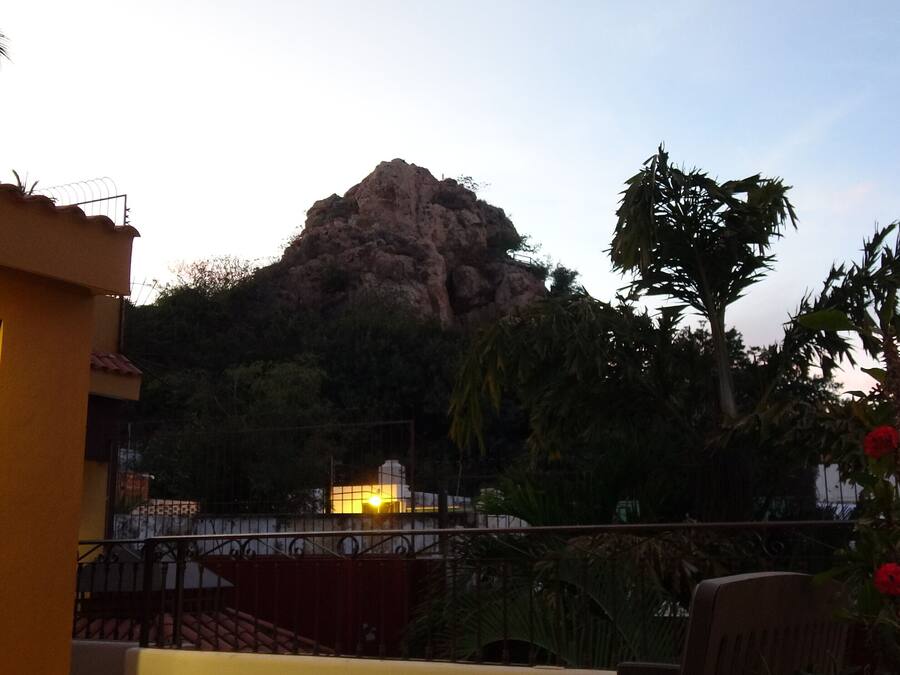 A balcony with a view of a rocky mountain, surrounded by greenery and a building with a yellow facade.