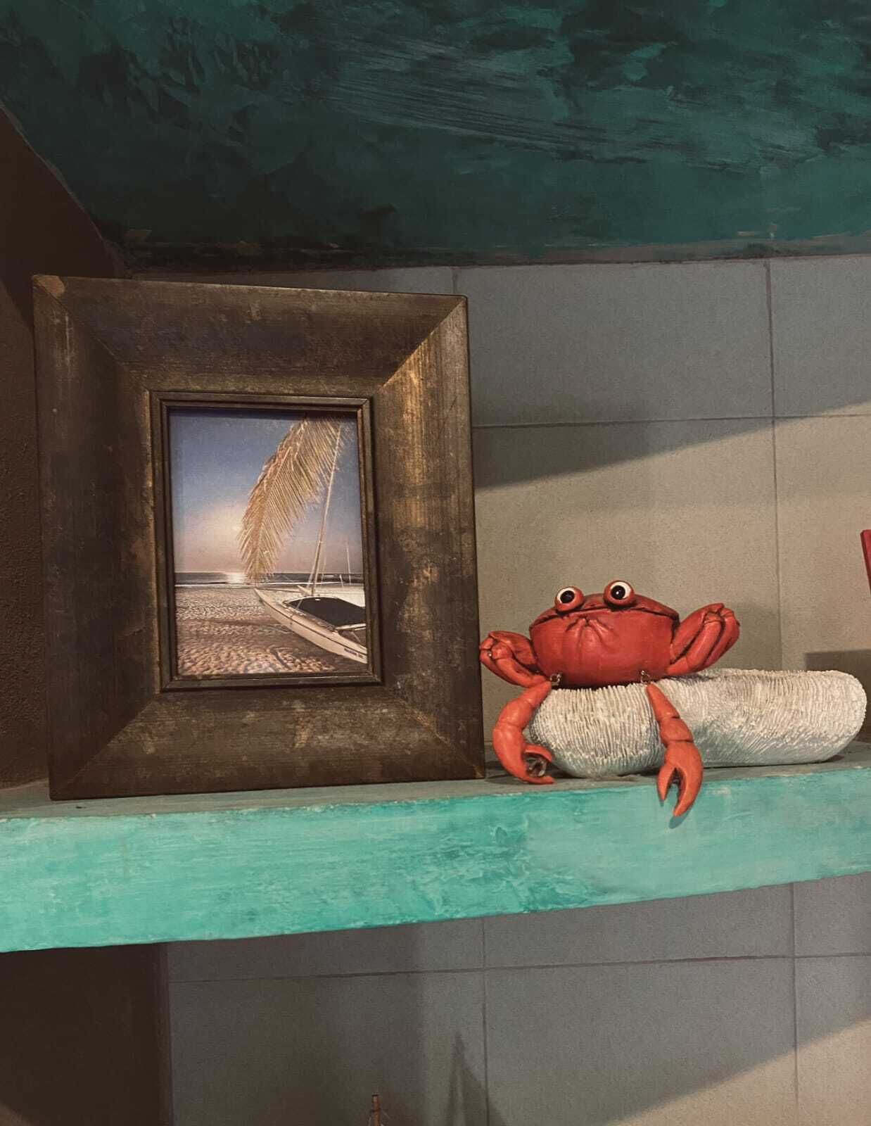 A framed picture of a beach scene on a shelf, a red crab figurine, and a teal-colored shelf against a tiled wall.