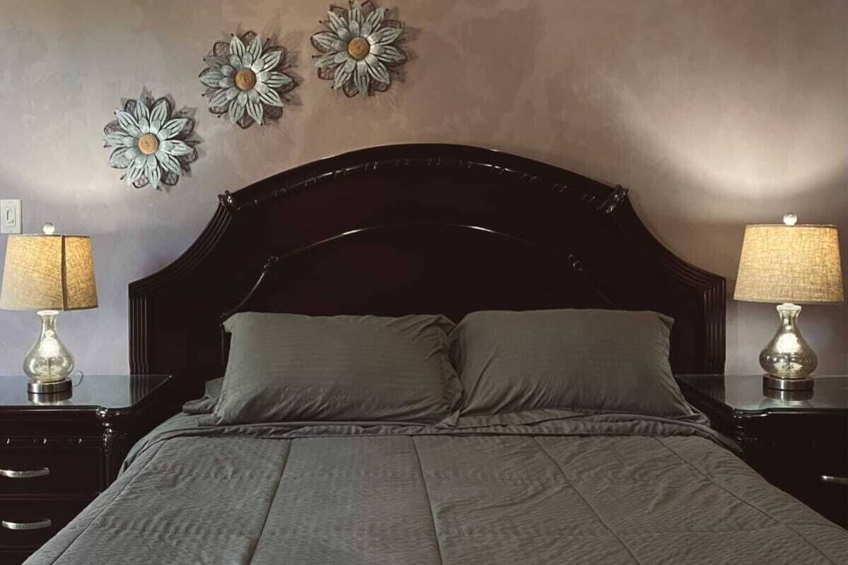 Quarto com cabeceira escura, duas mesas de cabeceira com luminárias e três flores decorativas na parede.