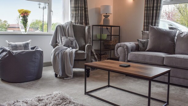 A modern living room with a grey sofa, a matching armchair, a wooden coffee table, and a grey pouf. There is a window with curtains, a lamp, and a vase with flowers on the windowsill.
