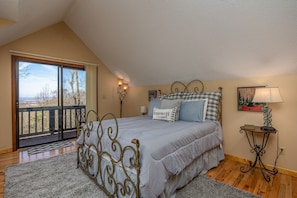 Relax in this cozy loft-style room with a large bed, wall-mounted lamps, and a sliding glass door leading to a balcony with a scenic view.