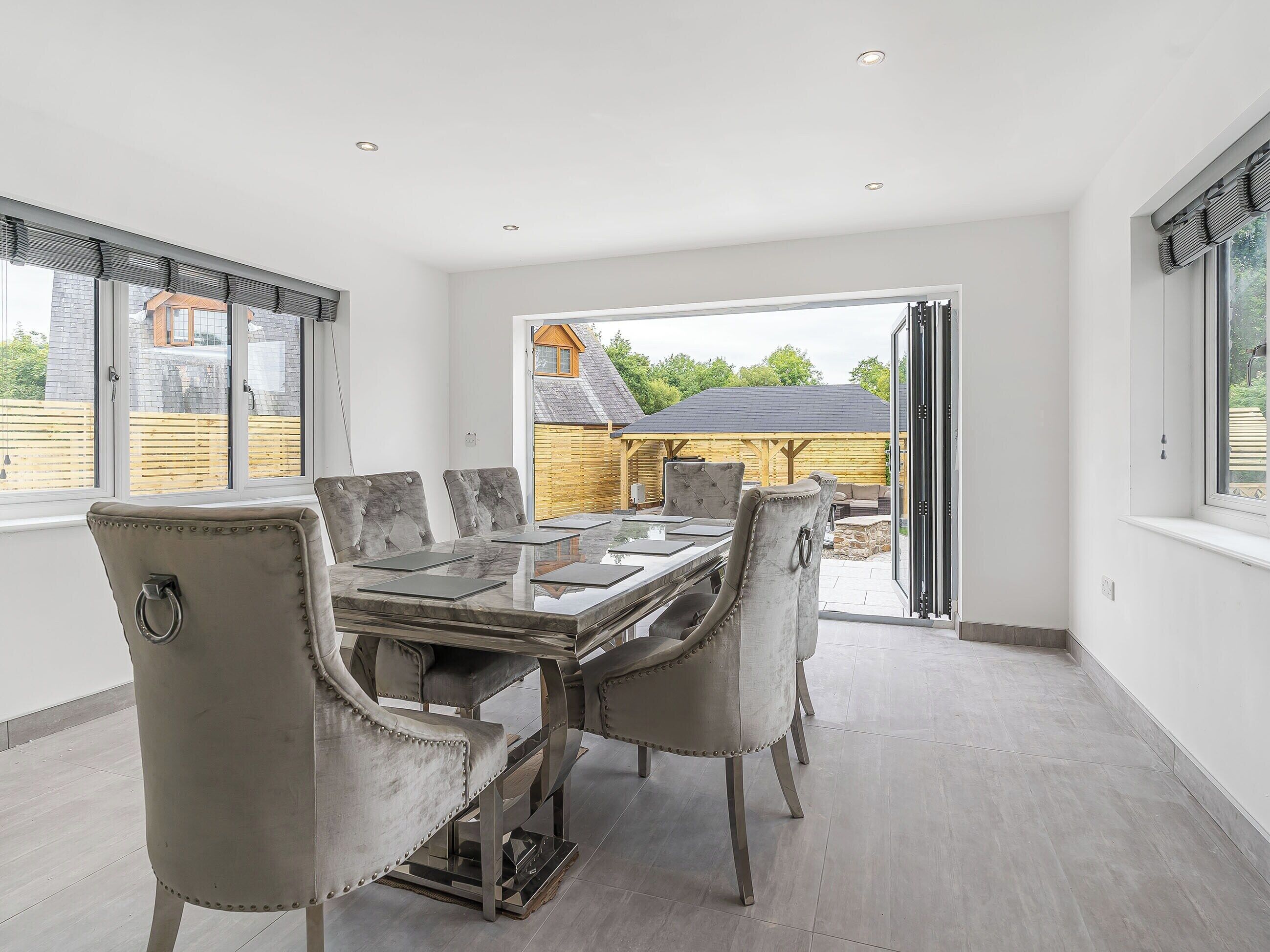 A spacious dining area with a large table and six upholstered chairs, a sliding glass door leading to an outdoor patio, and a view of a wooden deck and house.