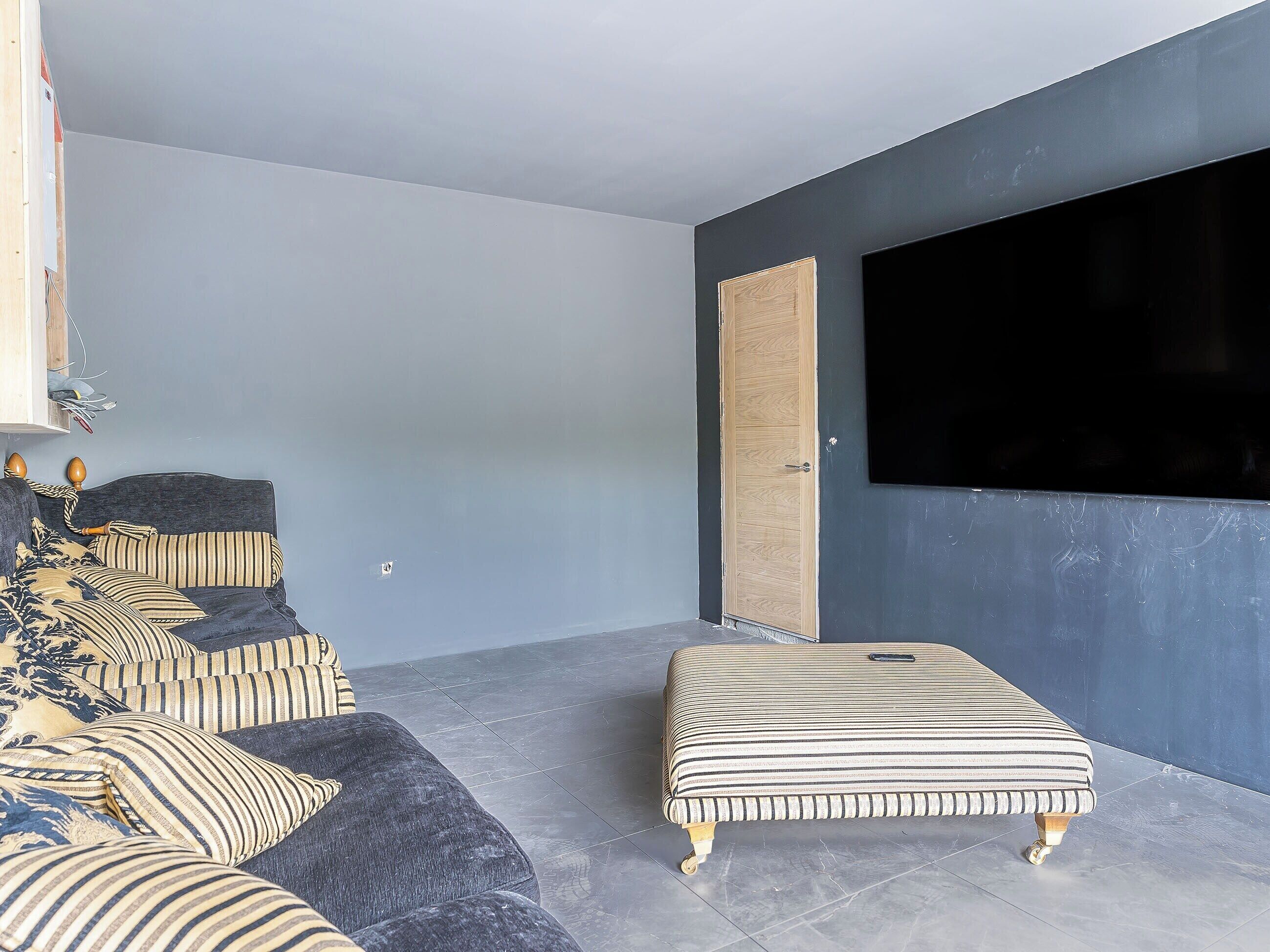Een moderne woonkamer met een donkere bank, gestreepte kussens, een ottoman met zebraprint, een houten deur en een grote televisie aan de muur.