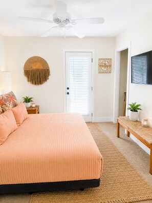 A bedroom with a bed, a ceiling fan, a door, a wall-mounted TV, and a bench with a plant.