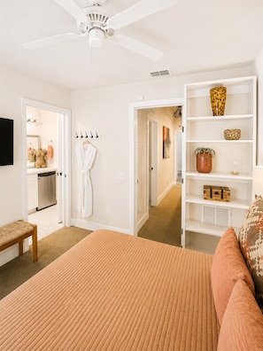 A bedroom with a bed, a white ceiling fan, a white wardrobe, a mirror, and a bathroom visible through an open door.