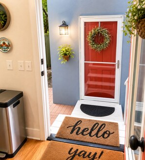 A red door with a wreath, a welcome mat, a potted plant, and a wall-mounted light fixture.