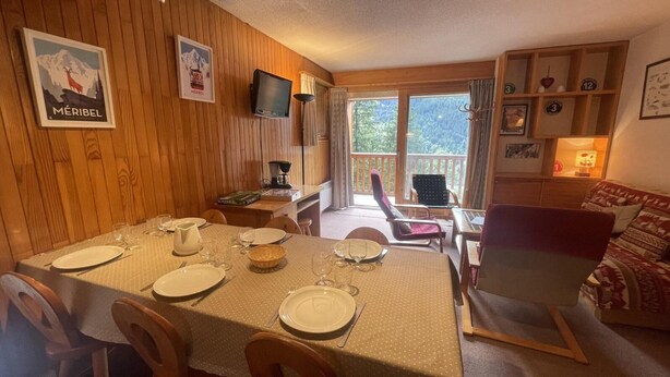En spiseplads med et træbord dækket til fire personer, et fjernsyn ophængt på væggen og en balkon med udsigt til bjergene.