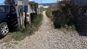 Un camino de grava que lleva a una playa, con un coche aparcado a la izquierda y una valla de madera a la derecha.