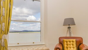A room with a view of a body of water, a window with curtains, a lamp, a brown leather armchair, and a beige ottoman. - Crow's Nest (Carnforth)