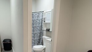 A narrow hallway leading to a bathroom with a toilet, a shower curtain, and a small shelf. A trash bin is visible on the left, and a purple chair is on the right. - Cozy studio just 5 min. Away from Coachella fest (Coachella)