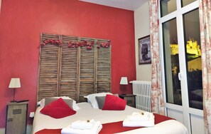 A hotel room with a red wall, a wooden shutter headboard, two beds with red and white bedding, a radiator, and a window with curtains.