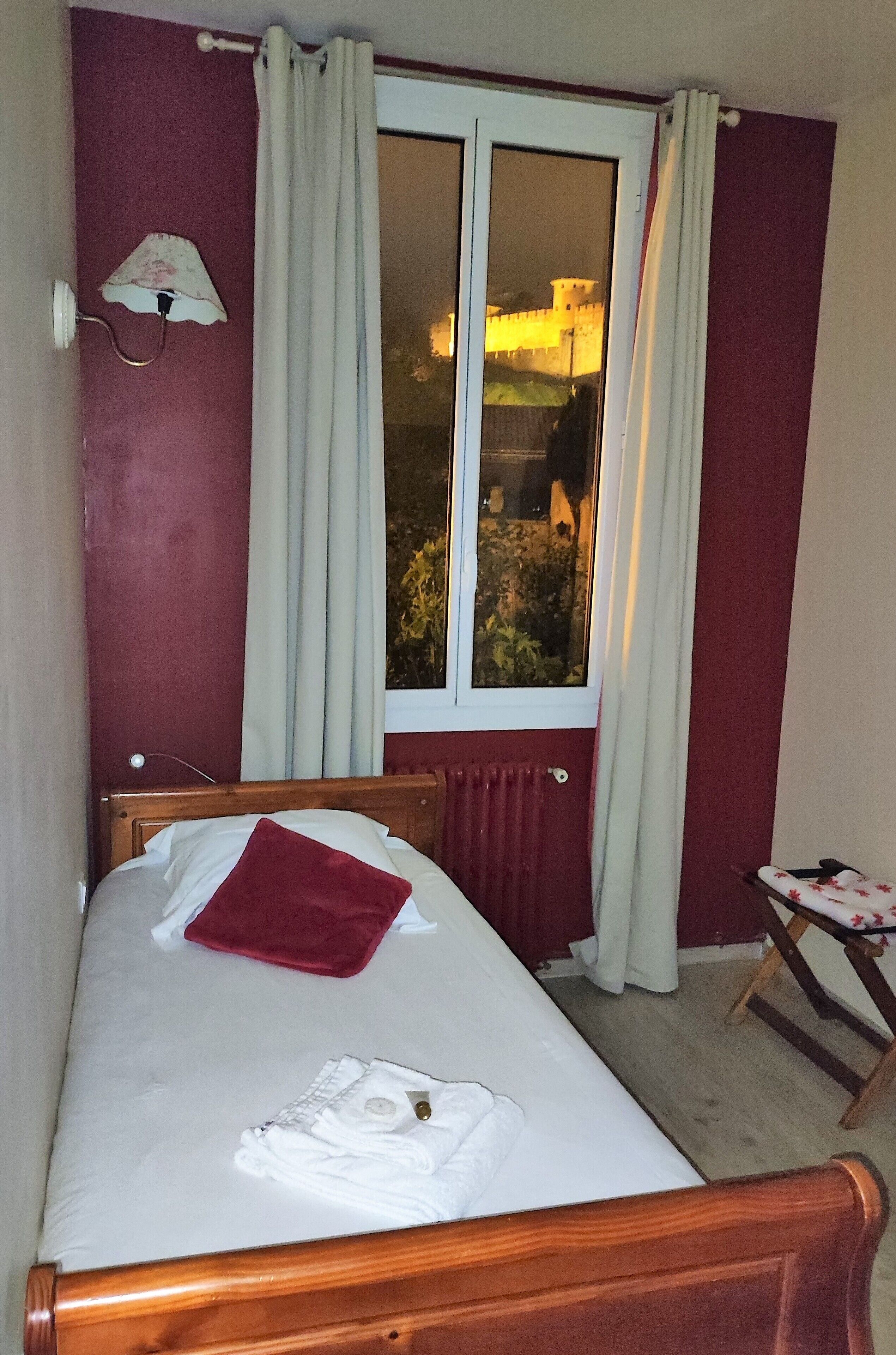 Quarto com cama, janela com cortinas, radiador e luminária fixada na parede.