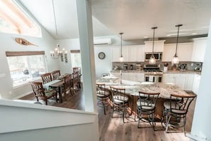 A modern kitchen with white cabinets, a large island with bar stools, pendant lighting, and a dining area with wooden chairs and a table. - Sunset Hideaway. Private 5 minute walk to lake.  (Cascade)