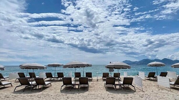 A beach with rows of sun loungers and umbrellas, a clear sky, and a view of the sea and mountains in the distance.