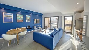 A modern living room with a blue sofa, a dining table with chairs, a TV, and a balcony with a view of buildings.