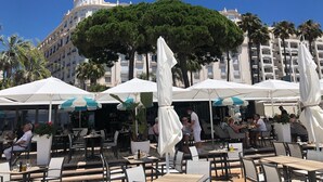An outdoor dining area with white umbrellas, tables, and chairs, with a large tree and a building in the background.