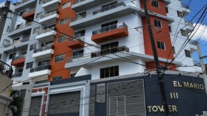 Un immeuble à plusieurs étages doté de balcons, une enseigne indiquant « TOUR EL MARIO », et des voitures garées devant.