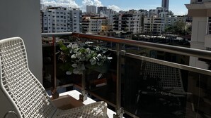 Un balcon agrémenté de chaises en osier blanc et d’une table ronde, d’où l’on peut admirer une vue panoramique sur la ville et ses différents bâtiments.