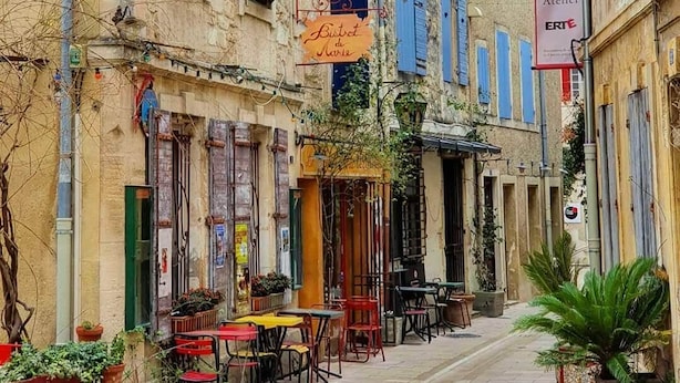 Eine enge Straße mit Gebäuden, die blaue Fensterläden und ein Schild für 'Galerie Lacaux Atelier ERTE' zeigen.