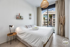 Un dormitorio moderno con una cama grande, una lámpara de noche, una silla de mimbre y vistas de edificios a través de una ventana.