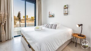 Ein Schlafzimmer mit einem großen Bett, einer Nachttischlampe und Blick auf Palmen durch ein Fenster.