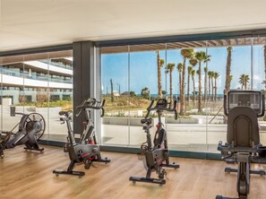 A gym with various exercise equipment, large windows offering a view of palm trees and a building. - Fari | Beach - terrace - pool (Torremolinos)